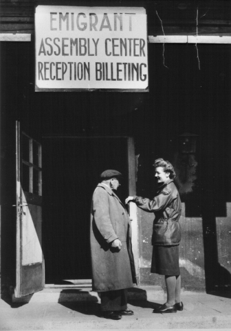 Black and white World War II era image of a man and woman, standing outside an open door, beneath a sign in all capital letters, which says "Emigrant assembly center reception billeting."